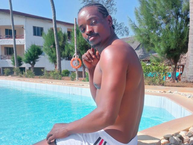 HoneyBadgers, shirtless with dark skin and a mustache, sitting by a blue pool, wearing white swim trunks, looking at the camera with a slight smile.