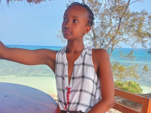 HoneyBadgers with dark brown skin, small tits, and a plaid sleeveless top, sitting at a beach table, arm raised, looking to the side, with blue ocean in background.