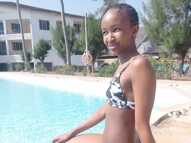HoneyBadgers with dark skin, small tits, and a black-and-white bikini, sitting by a blue pool, smiling, with curly hair and small earrings, in a sunny outdoor setting.