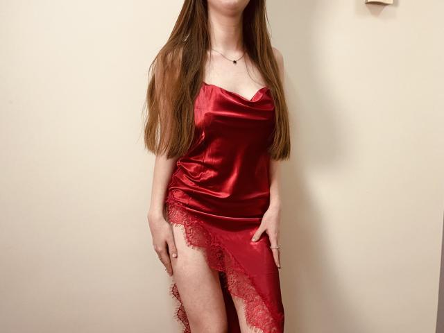 Nesie, com cabelos castanhos longos, vestindo um vestido de seda vermelho, mostrando uma coxa com renda, sua mão sobre a coxa, seios pequenos, pele clara e um colar..
