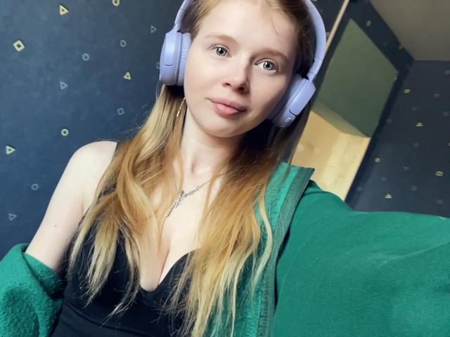 MeeBoggio has blonde hair, light skin, and small tits, wearing a green zip-up and black tank top, with purple headphones, smiling slightly, in a selfie.