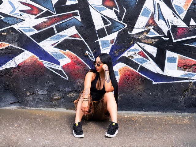 ChloeMoretti, squatting against a graffiti-covered wall, has long black hair, wearing sunglasses, a black tank top, and brown shorts. She has tattoos on her left forearm and right bicep. She's wearing black sneakers with yellow accents. The graffiti features abstract geometric patterns in black, white, and blue, with red highlights. Her right arm rests on her knee, while her left hand touches her face. The concrete ground is visible.