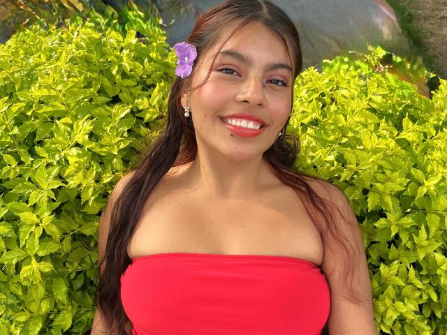 A photograph of AlyssaGreen with long brown hair, a purple flower in her hair, and wearing a red strapless top. She has a bright smile with red lipstick, small earrings, and light brown skin. She's surrounded by bright green shrubs. The image focuses on her upper body and face, highlighting her cheerful expression and natural beauty. The colors are vibrant, with the red top contrasting sharply against the green foliage.