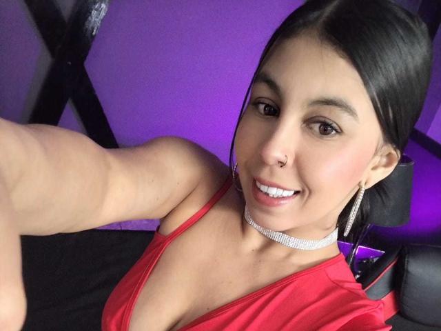 NenaBlack has medium-length black hair, brown eyes, and a nose piercing. She wears a red sleeveless top, a silver choker necklace, and silver hoop earrings. Her smile reveals white teeth. She extends her arm for a selfie, showing a slightly muscular arm. The purple background and black cross-shaped object are blurred. The image focuses on her upper body and face. The lighting highlights her skin tone and makeup.