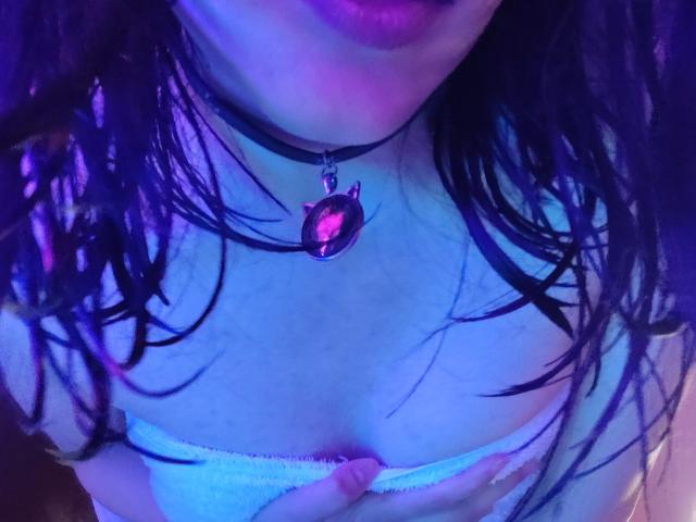 IssisDiosa with wet black hair, purple-blue lit skin, wearing a black choker with an oval pendant, and pulling down white lingerie to expose her small tits.
