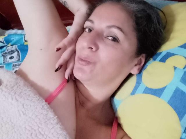 MadameScarlett with pouty lips, dark nails, pink strap, armpit raised, lying on yellow and blue patterned pillow, looking at camera. She has a tattoo on her arm.