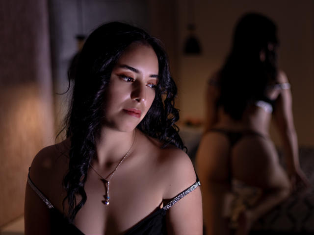 MartinaRey with curly black hair, looking down, wearing a black top with off-shoulder straps, necklace, and thong, showing her ass in the mirror's reflection.