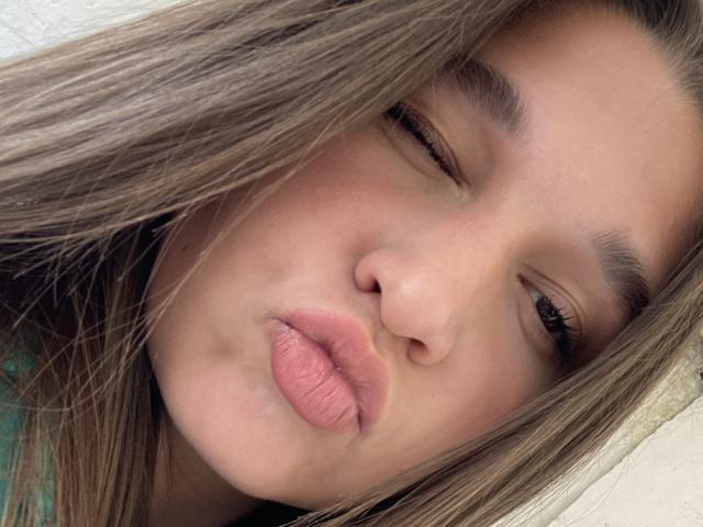 Close-up photo of MissKuromi's face. She has light brown, slightly wavy hair that partially covers her right eye. Her left eye is closed, and she's puckering her lips in a playful kiss. She has a fair skin tone, visible freckles, and subtle makeup. Her eyebrows are dark and slightly arched. The texture of her hair is smooth, and the light in the image is soft, creating gentle shadows. The focus is on her facial expression and details of her skin and hair.