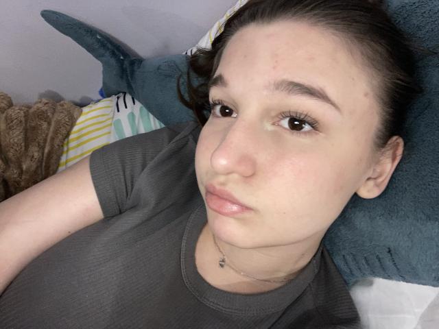 Close-up selfie of MissKuromi with fair skin and dark brown hair in a messy bun. She has brown eyes, small nose, and slightly parted pink lips. She's wearing a gray t-shirt and a thin necklace. She's lying on a teal pillow and a striped pillow. The texture of the pillow is soft and fuzzy. Her face has small blemishes. The light is even, highlighting her features clearly. The image has a casual, relaxed vibe.