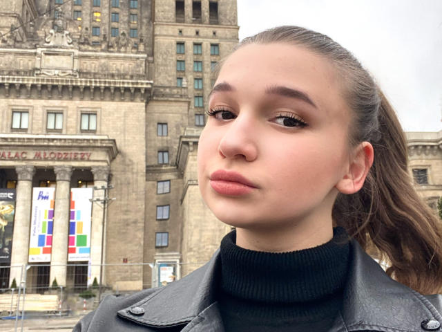 Portrait of MissKuromi with a serious expression. She has fair skin, brown eyes, and long brown hair in a ponytail. She's wearing a black turtleneck and a black leather jacket. Her eyebrows are slightly raised. The background features an ornate, stone building with multiple windows and a colorful banner. The building has a classical architectural style. The image is a photograph. Her face is centered, with a slight upward angle.