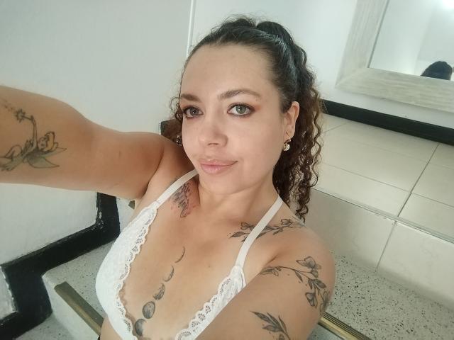 Selfie of RubbyStrongX with curly brown hair in a high ponytail, wearing a white lace bra. RubbyStrongX has light brown skin, green eyes, and small earrings. Tattoos on both arms and chest, including floral and geometric designs. RubbyStrongX is smiling slightly, looking at the camera. The image is clear, with natural light. The background has white tiles and a black metal pipe. RubbyStrongX's right arm is extended, taking the selfie. The photo is focused on RubbyStrongX's upper body.