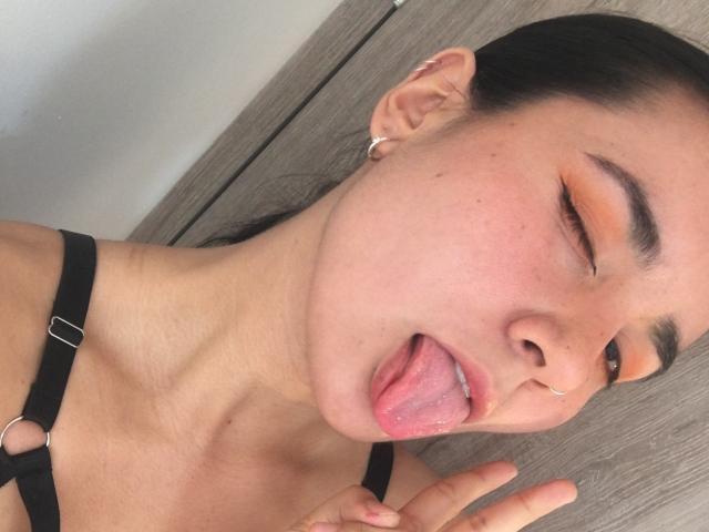 Natii69 with dark hair, pinkish-orange eye makeup, nose ring, tongue out, and black strap top, making a peace sign. Her skin is light, and she's winking.