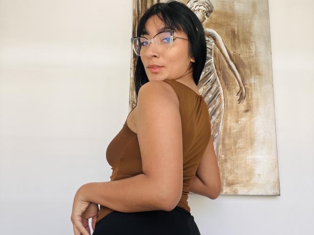 AmelineRousseau stands with her back to the camera, head turned to the right. She has black hair, glasses, and wears a brown sleeveless top with black pants. Her right arm is slightly raised. She has light brown skin and a confident expression. Behind her, a beige painting of a skeletal figure is visible. The painting is textured with a rough, brush-stroke finish. AmelineRousseau's glasses have a thin, clear frame. Her facial features include a slight smile and dark eyebrows. The image is clear and well-lit.