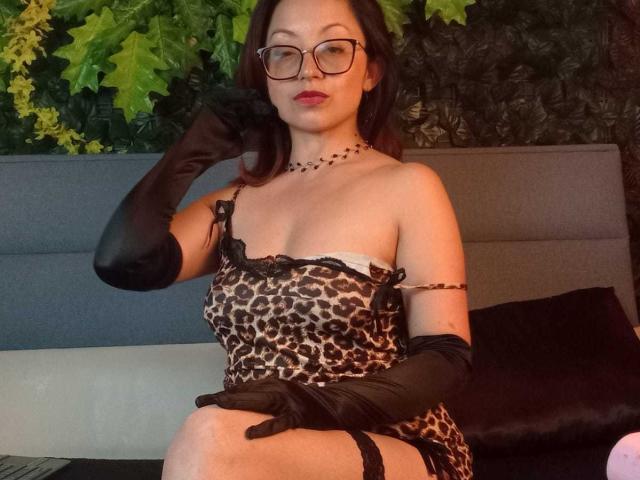 AngelinaBlanche with glasses, wearing a leopard print dress, black gloves, and a necklace, sitting on a bed, showing her tits and ass, with a confident, sexy look.