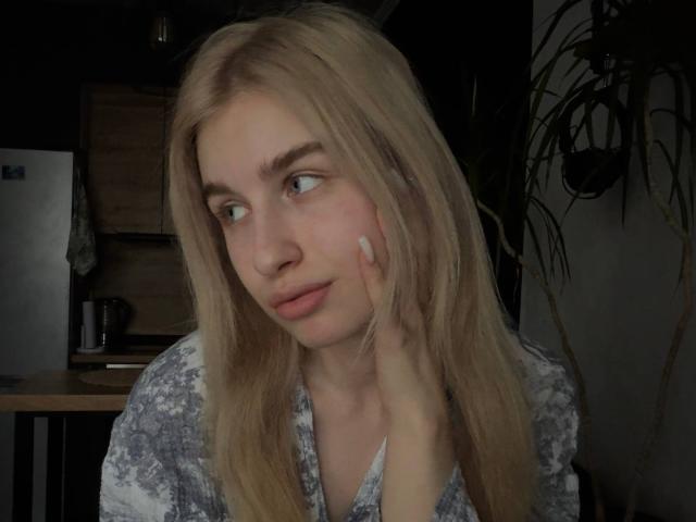 Blond, fair-skinned KathrineFleak with blue eyes and pink lips, touching her cheek with a hand, wearing a gray and white floral shirt, looking to the side.