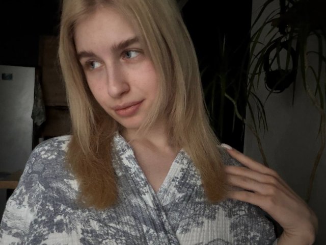 KathrineFleak has blonde hair, blue eyes, and a light skin tone. She's wearing a gray and white patterned shirt, with one hand near her neck, looking slightly to the side.