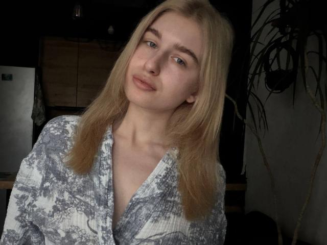 KathrineFleak with blonde hair, blue eyes, and a slight smile, wearing a grey and white patterned shirt, has a cute face and small tits. She looks relaxed.