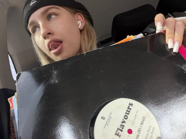KathrineFleak with blonde hair, black beanie, and white earbuds, sticking out tongue, holding black vinyl record, wearing white nail polish, inside car.