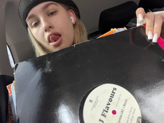 KathrineFleak with blonde hair, black beanie, and white earbuds, tongue out, holding a black vinyl record, pink nails, in a car, looking at the camera.