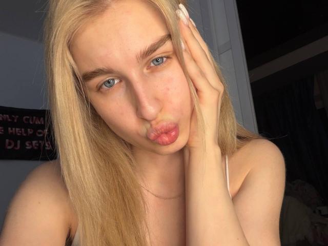 KathrineFleak has blonde hair, blue eyes, and pouts her lips while touching her face with her right hand, showing off her white nails. She's topless with a fair skin tone.