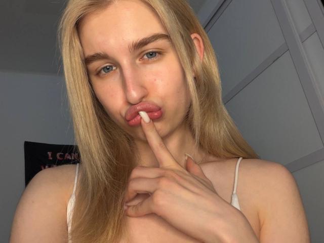 KathrineFleak with blonde hair, pouty pink lips, and blue eyes, sucking on her index finger, wearing a white tank top, in a well-lit room.