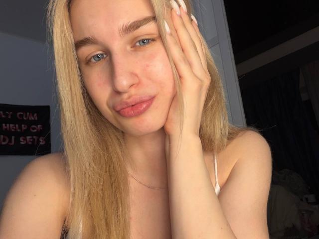 KathrineFleak with blonde hair, blue eyes, and pink lips, pouting slightly, touching her face with a hand, wearing a white strap, in a sunlit room.