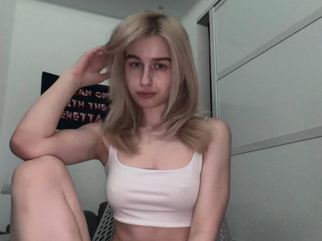 KathrineFleak with blonde hair, white tank top, small tits, sitting, one knee up, hand in hair, slight smile, pale skin, in a simple room.