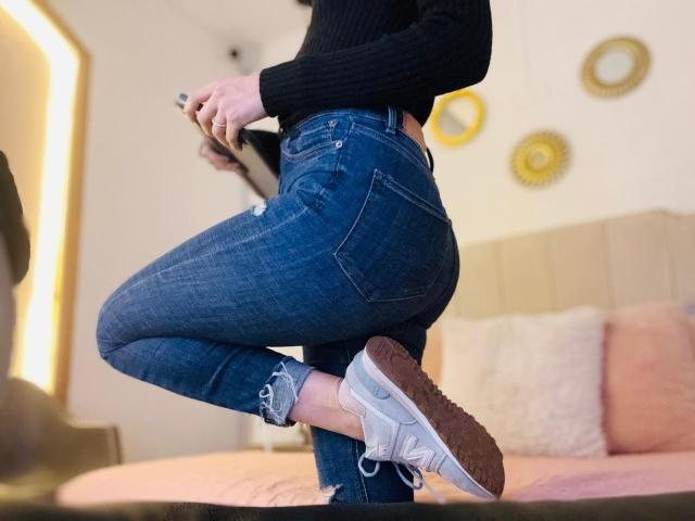 AdharaCooperr stands in a room, captured from a side angle. She wears tight, blue, ripped jeans that highlight her curvy, toned buttocks and a black, long-sleeved, rib-knit top. Her right leg is bent at the knee, foot resting on a surface, showing white, lace-up, athletic sneakers with brown soles. She holds a phone in her right hand. The image focuses on her lower body and jeans, with a blurred, beige, cushioned sofa in the background. The composition is dynamic, emphasizing her pose and denim texture.