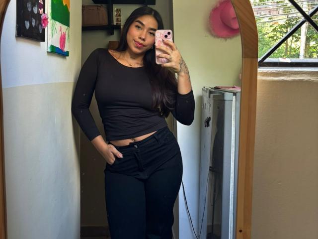 Saalome69 with long brown hair, wearing a black long-sleeve top and black pants, holding a pink phone, showing a tattoo on her right hand, and her hand in her pants.