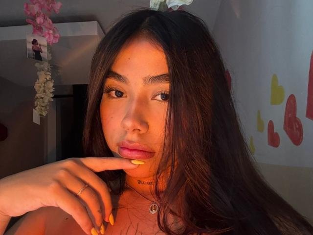 Saalome69 with long brown hair, orange light, yellow nails, finger on lips, pink lipstick, choker, ring, and a small nose piercing, looking at camera.