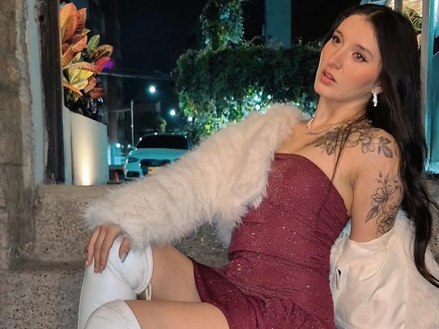 ZoeAvancini with long dark hair, wearing a sparkly red dress, white fur jacket, and white boots, showing tattoos on her right arm and thigh, sitting at night.