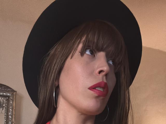 SeleneBrigth has brown bangs, red lipstick, and wears a black hat with silver hoop earrings, looking up with a slightly open mouth. Her skin is fair.