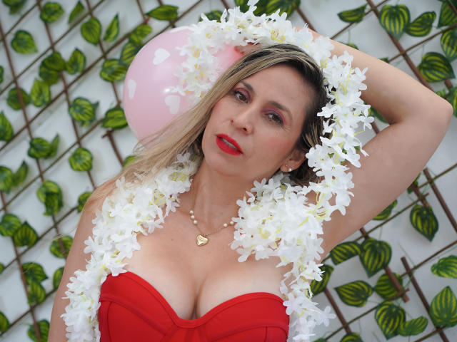 DianaPearlsMilf in a bright red strapless dress, her blonde hair partially covering her face, wears white floral garlands on her head and chest. She has red lipstick and a pearl necklace with a small heart pendant. Her right arm is raised, hand behind her head. A pink balloon is visible behind her. Her chest is prominent with cleavage. The image has a casual, celebratory feel with a green and white lattice pattern in the background. She has a fair skin tone. The composition is centered on DianaPearlsMilf.
