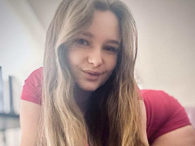 MagikPeachh with long, blonde hair, wearing a red shirt, slightly pouting, smiling softly at the camera, leaning forward, looking cute and sexy.