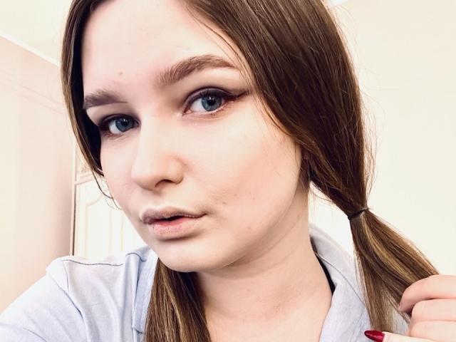 MagikPeachh has light skin, blue eyes, and brown hair in a ponytail, wearing a light gray shirt, with red nails, and a slight pout, looking at the camera.