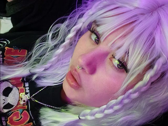 MoonBluee with white-purple hair, braided, nose piercing, green eyes, glossy lips, black shirt, skull pattern, purple lighting. Her face is slightly tilted, looking at the camera.