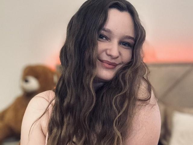 MagikPeachh with long wavy brown hair, smiling, showing her tits, freckles, and subtle makeup, in a soft bedroom setting with a blurred teddy bear in the background.