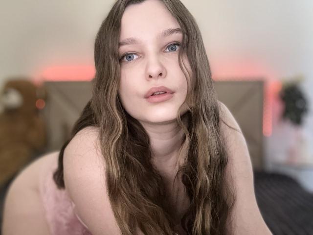 MagikPeachh with long brown wavy hair, pale skin, blue eyes, and pink lingerie, leaning forward, looking into camera, slightly parted lips, soft expression, in bedroom setting.
