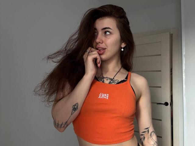 ClarindaWiatr with long brown hair, pouty lips, wearing an orange sports bra, showing tattoos on arms, looking to the side, finger near mouth, small earrings.
