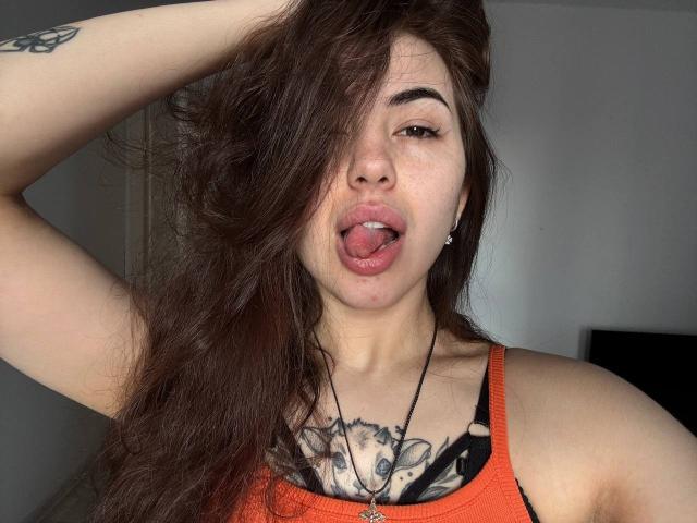 ClarindaWiatr with long brown hair, tongue out, wearing an orange tank top, showing tattoos on her chest and arm, hand in hair, looking at camera.