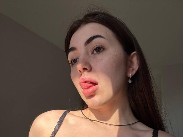 ClarindaWiatr with pouty pink lips, brown eyes, and long dark hair, wearing a black strap, showing a hint of her tits, with small earrings and a thin necklace.