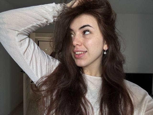 ClarindaWiatr with messy dark brown hair, white long-sleeve shirt, smiling, hand in hair, blue earrings, light skin, small nose, and subtle freckles, looking to the side.