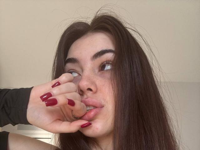 ClarindaWiatr with long dark brown hair, freckles, and red nails, sucking on her index finger, looking to the side, wearing a black sleeve, and a nose ring.