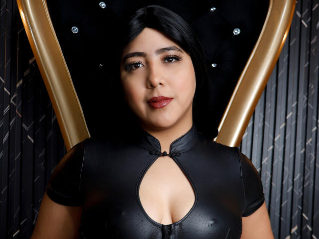 A close-up of SamyWalker, a woman with long black hair and medium brown skin, wearing a tight black leather outfit with a keyhole cutout that reveals her cleavage. She has dark brown eyes, full red lips, and a confident expression. The background features a black, diamond-patterned wall with golden, V-shaped accents. Her outfit is shiny and form-fitting, highlighting her curvy physique. The lighting emphasizes her facial features and outfit texture.