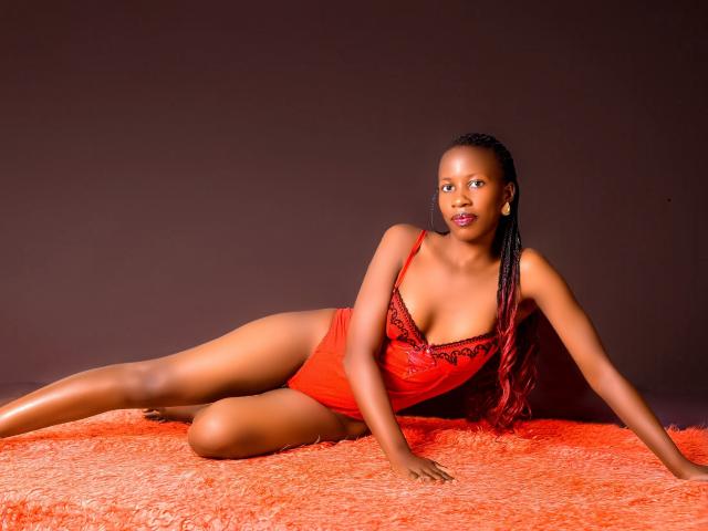 KellyAlexa is lying on an orange, textured rug. She's wearing a red, lace-trimmed one-piece. KellyAlexa has dark skin and long, braided hair. She's looking to the right with a neutral expression. Her right arm is extended, supporting her upper body, while her left arm is bent at the elbow. She's wearing yellow earrings. The rug and her outfit create a strong contrast with her skin tone. The lighting is even, highlighting her figure and the texture of the rug.