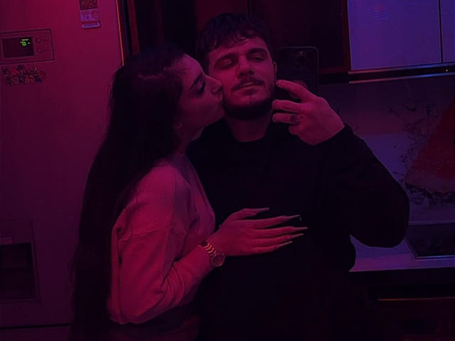HardAnal69 with long dark hair and tits, kissing HardAnal69 with short dark hair and a beard, who's taking a selfie. Red and blue lighting, HardAnal69's hand on chest.