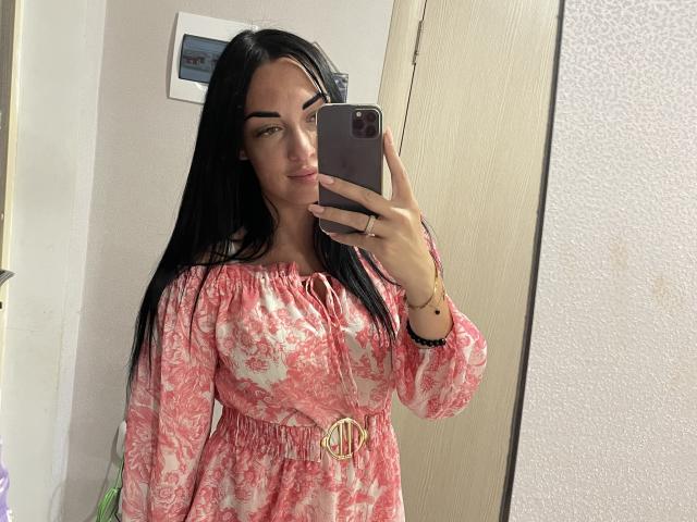 Xioni with long black hair, pink floral dress, and gold belt, taking a mirror selfie with her phone. She has a confident smile and a ring on her left hand.