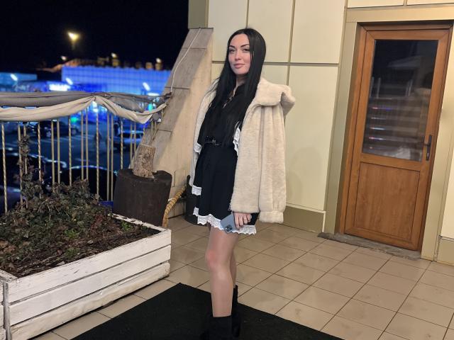 Xioni has long black hair, pale skin, and wears a black dress with white lace, a beige fur coat, and black boots. Standing on a tiled balcony at night.
