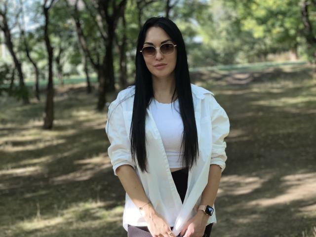 A photograph of Xioni standing in a park. She has long black hair, wears round sunglasses, and looks directly at the camera. She's dressed in a white, slightly oversized button-up shirt with sleeves rolled up, and a white crop top. She also wears dark pants. Her hands are clasped in front of her. She has a watch on her left wrist. The image has a natural, relaxed composition with a slight blur in the background, emphasizing Xioni as the central focus.