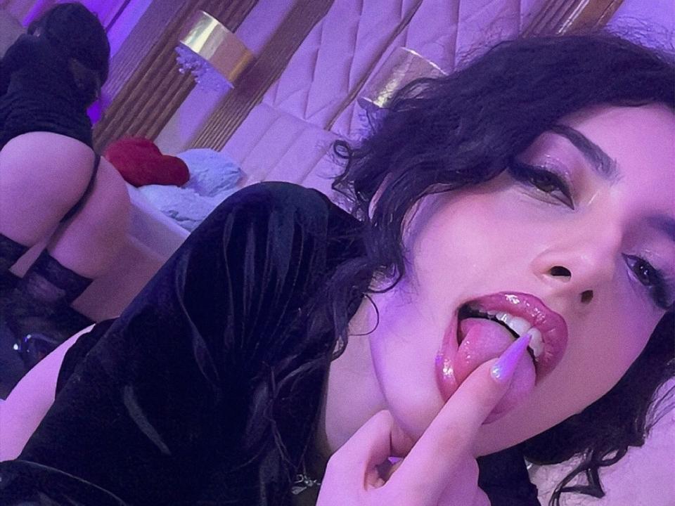 HeidiCandyx with black hair, glossy lips, and purple eyeshadow, tongue out, finger in mouth, showing off her glossy pink tongue and black lingerie, her ass in the background.